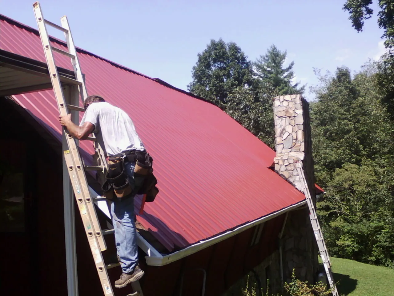 Roofer on a ladder beside a red metal roof and stone chimney — Soffit & Fascia Repair by Roofing Brothers in Havana